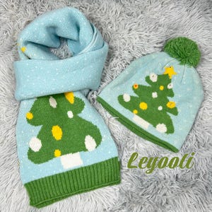 May include: A light blue knit scarf and beanie set. The scarf and hat feature a green Christmas tree design with yellow and white accents. The hat has a green pom-pom on top. The set is on a fluffy, gray surface.