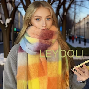 Puede incluir: Una bufanda de cuadros coloridos con tonos de rosa, amarillo, naranja y azul. La bufanda está hecha de un material suave y peludo y la lleva puesta una persona con el pelo largo y rubio. El texto "LEYDOLI" es visible en la imagen.