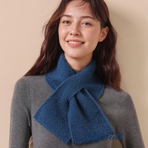 May include: A deep blue knitted scarf with a textured, pebbled surface. The scarf features a pull-through design where one end passes through a slit in the other, creating a secure, layered loop around the neck. It is worn over a grey ribbed sweater.