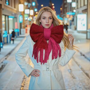 Op de afbeelding: Een vrouw draagt een witte jas en een grote, rode strikvormige sjaal. De sjaal heeft een donkerrode strik en een lichtere rode franje. De achtergrond is een stadsstraat 's nachts met vage lichten.