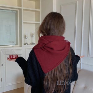 May include: A deep red knitted scarf with a triangular shape and a single button closure on the side. The scarf features a textured ribbed knit pattern and is worn around the neck, draping over the shoulders. The fabric is a solid dark burgundy colour.