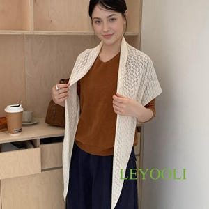 Puede incluir: Una mujer lleva un chal crema de tejido abierto sobre una blusa marrón y pantalones azul marino. El chal tiene un patrón geométrico. La palabra "LEYOOLI" está impresa en verde en la parte inferior derecha de la imagen. El fondo incluye estanterías de madera.