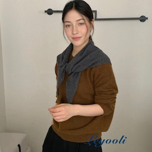 Puede incluir: Una mujer con un suéter marrón y una bufanda gris alrededor del cuello. El suéter tiene mangas largas y se combina con pantalones negros. El fondo es de color neutro.
