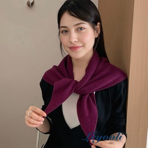 May include: A dark purple scarf is worn over a black top, knotted at the front. The scarf's ends form a triangle shape. The model is wearing a white undershirt. The scarf appears to be made of a soft, knitted fabric. The outfit has a casual, yet stylish appearance.