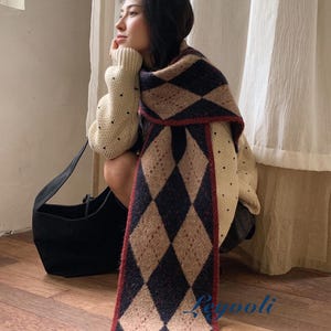 May include: A long knit scarf featuring a repeating argyle pattern in dark navy blue and tan, finished with a thin red border. The scarf is paired with a cream-colored sweater with small black dots and a black shoulder bag.