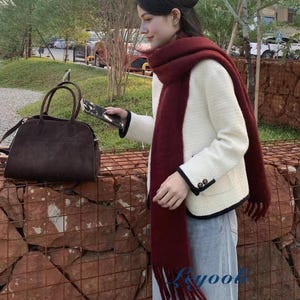 May include: A woman wearing a white jacket with black trim, a burgundy scarf, and blue jeans. A brown handbag with a top handle and a crossbody strap is on the left. The background includes trees and a brick wall.
