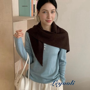 May include: A woman wearing a light blue long-sleeve shirt with a brown scarf. She is also wearing white pants and carrying a white handbag. The word "Gravity" is visible on a book on a shelf.