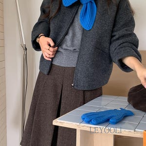 以下が含まれることがあります： チャコールグレーのウール混ジャケットにグレーのセーター、ブラウンのプリーツスカート、鮮やかなブルーのリブニットスカーフを着用。LEYOOLIという文字がプリントされたタイル状の表面に、お揃いのブルーのニット手袋が置かれています。