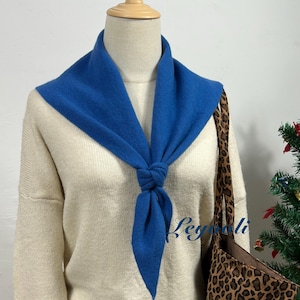 Pode incluir: Um cachecol azul amarrado, usado sobre um suéter creme. O cachecol é azul vibrante e contrasta com os tons neutros do suéter. Uma sacola tote com estampa de leopardo é visível ao fundo.