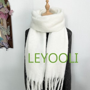 May include: A soft, white scarf with a fringed edge and the text "LEYOOLI" in green. The scarf is draped over a dark gray garment, creating a contrast. The scarf appears to be made of a fluffy, warm material.