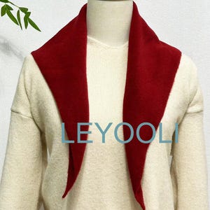 May include: A red fleece scarf with a pointed collar. The scarf is worn over a cream-colored sweater. The word "LEYOOLI" is printed in blue on the scarf.