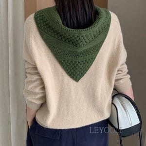 Pode incluir: Um suéter bege de manga comprida com um capuz triangular de crochê verde-escuro preso ao decote. O suéter é combinado com calças azul-escuras e uma bolsa branca e preta. O texto LEYOOLI é visível no canto inferior direito.