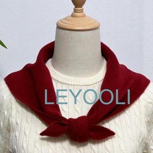 May include: A red knit scarf tied in a bow around a white sweater. The scarf is made of a soft, fuzzy material. The text "LEYOOLI" is printed on the scarf.