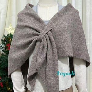May include: A taupe-colored shawl with a knotted design. The shawl is draped over a mannequin, showcasing its shape and texture. The garment appears to be made of a soft, knitted material, suitable for cooler weather. The shawl is a versatile accessory.