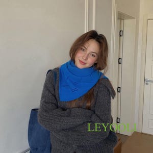 May include: A woman wearing a gray cardigan and a bright blue scarf. The scarf has a unique cutout design. The cardigan is buttoned and appears to be made of a soft, textured material. The text "LEYCOLL" is visible on the image.