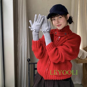 May include: A red knit sweater with a high collar and a zipper, paired with a brown skirt. The outfit includes gray gloves with bow details. A navy blue cap is also worn.