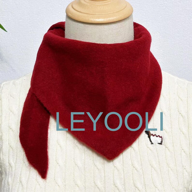 May include: A red fleece neck warmer with a triangular shape. The neck warmer is on a white sweater with a cable knit pattern. The text "LEYOOLI" is visible on the neck warmer.