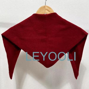 May include: A burgundy red triangular scarf hanging on a wooden hanger. The scarf is made of a soft, fuzzy material. The word "LEYOOLI" is printed in white on the scarf.