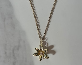 Puede incluir: Un collar dorado con una delicada cadena y un colgante de flor. La flor tiene seis pétalos y una pequeña gema rosa en el centro. El collar se muestra sobre una superficie de mármol blanco y gris.