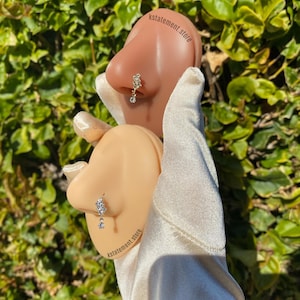 May include: Two nose rings are shown, one with a floral design and dangling gem, the other with a single dangling gem. The jewelry is displayed on a tan-coloured nose form, held by a gloved hand. The background is a lush green bush. The text "kstatement.store" is visible.