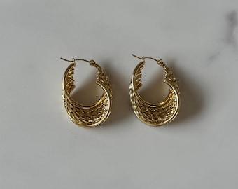 May include: A pair of gold-colored hoop earrings. The earrings have a woven design and a hinged closure. The earrings are oval-shaped and are displayed on a white and gray surface. The earrings are a jewelry accessory.