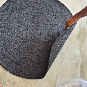 Puede incluir: Una alfombra redonda de color gris carbón oscuro con una construcción trenzada y texturizada. La alfombra tiene forma circular y muestra un patrón tejido consistente en toda su superficie, con un borde levantado que revela el reverso.
