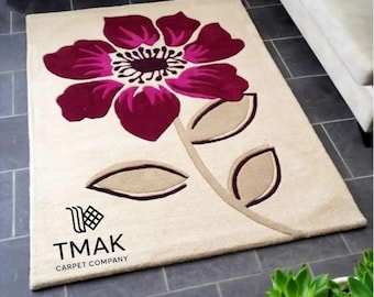 Könnte beinhalten: Beiger rechteckiger Teppich mit großem Blumendesign. Die Blume hat tiefmagentafarbene Blütenblätter und ein schwarz-weißes Zentrum. Der Stiel und die Blätter sind beige mit braunen Umrissen. Der Teppich trägt den Text "TMAK CARPET COMPANY" in Schwarz.