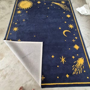 May include: A rectangular area rug with a deep navy blue background and a yellow celestial pattern featuring a sun, a crescent moon, stars, and two comets. The rug has a yellow border and one corner is folded back to reveal a light grey backing.