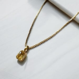 Puede incluir: Un collar de cadena dorada con un pequeño colgante rectangular con una piedra preciosa amarilla.