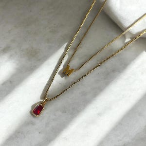 Peut inclure: Un collier en chaîne dorée avec un pendentif en pierre rouge et un charme en lettre M dorée.