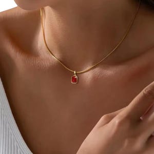 Könnte beinhalten: Eine goldfarbene Halskette mit einem kleinen, rechteckigen, roten Anhänger. Der Anhänger ist in einen goldenen Rahmen gefasst und hängt an einer zarten Kette. Die Halskette wird um den Hals getragen.