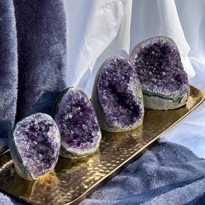 May include: Five amethyst crystal geodes of varying sizes on a gold tray. The geodes are hollow and filled with purple crystals.