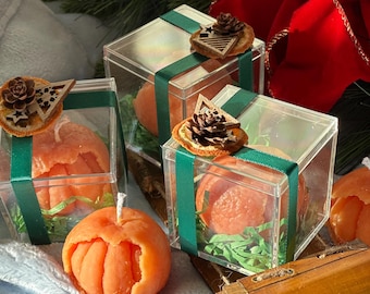 May include: Four orange scented candles in clear boxes with green ribbon and a wooden Christmas tree ornament. The boxes are sitting on a wooden sled with a brown background.