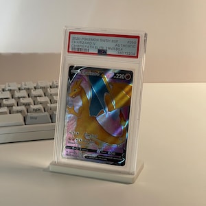 May include: A graded Charizard V Pokémon card in a protective case. The card features a vibrant orange and blue dragon illustration with the text "Charizard V" and "220". The case is displayed on a white stand.