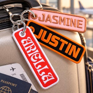 May include: Three rectangular embroidered luggage tags attached to a silver metal ring. One tag is light pink with the name JASMINE in brown, one is black with the name JUSTIN in orange, and one is red with the name ARIELLA in white embroidery.