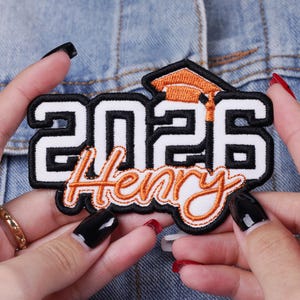 May include: Embroidered patch with the text "2026 Henry" in white, black, and orange. The patch features a graduation cap design. The patch is held by hands against a denim background.