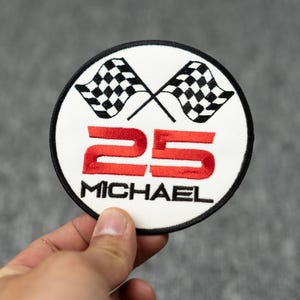 May include: White round embroidered patch with two black and white checkered flags crossed behind the red number 25 and the name "MICHAEL" in black.