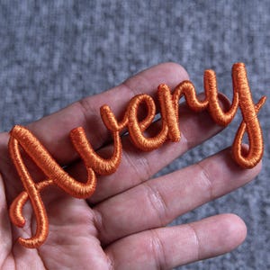May include: Orange embroidered patch spelling the name "Avery" in a cursive font. The patch is made of a textured thread and is displayed against a gray background. The letters are connected and have a flowing design.