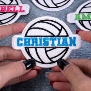 May include: Embroidered volleyball patch with the name "CHRISTIAN" in blue letters on a white background. The patch is round with a black outline and a volleyball design. Other patches with names are visible.