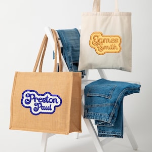 May include: Two tote bags with personalized embroidered names. The bag on the left is a brown jute tote bag with the name "Preston Paul" embroidered in blue. The bag on the right is a white canvas tote bag with the name "James Smith" embroidered in yellow.