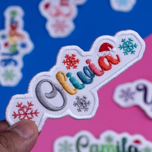 May include: Embroidered patch with the name "Olivia" in colorful thread, featuring a Santa hat and snowflakes. The patch is white with a decorative border. Other patches are visible in the background.