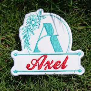 May include: White embroidered patch with the name "Axel" in red script. The letter "A" is in teal, surrounded by teal floral embroidery. The patch is on a background of green grass.
