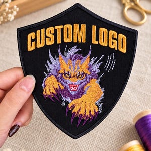 May include: A shield-shaped black embroidered patch featuring the yellow text CUSTOM LOGO at the top. Below the text is a detailed embroidered graphic of a snarling creature with purple and yellow fur, sharp claws, and white scratch marks.