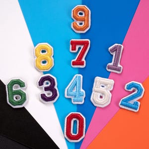 May include: A collection of embroidered fabric number patches, each with a white border. The numbers are in various colors, including orange, yellow, red, purple, blue, green, and white. The patches are arranged on a colorful background.