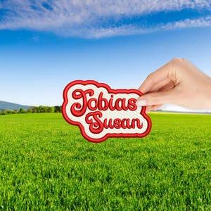 May include: A white patch with red embroidered text that reads "Tobias Susan" against a green grassy field and a blue sky.