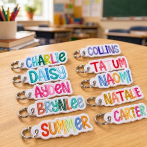 May include: A collection of ten white fabric keychains featuring embroidered names in various bright colors including red, orange, yellow, green, blue, and pink. Each rectangular keychain has a silver metal split ring attached to one end.