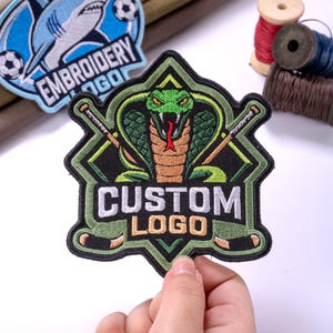 May include: A custom embroidered patch featuring a green cobra with a red tongue, positioned behind two crossed hockey sticks. The design includes the text CUSTOM LOGO in white and gold lettering, set against a black and olive green shield background.