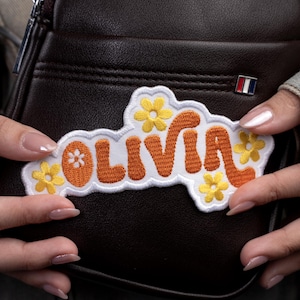 May include: Embroidered patch with the name "Olivia" in orange, outlined in white, and decorated with yellow flowers. The patch is held against a brown leather bag, showcasing a small tricolor pin.