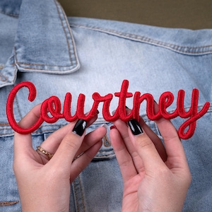 May include: A red embroidered patch with the name "Courtney" in a cursive font. The patch is held in front of a light blue denim jacket. The name is in a bold, eye-catching script.