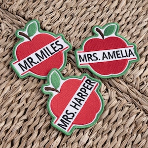 May include: Three embroidered apple-shaped patches with green leaves and stems. Each patch has a white banner with a name: "MR. MILES", "MRS. AMELIA", and "MRS. HARPER". The apples are red with green and white trim.